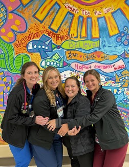 four residents, wearing scrubs and hugging, pose in front of a mural